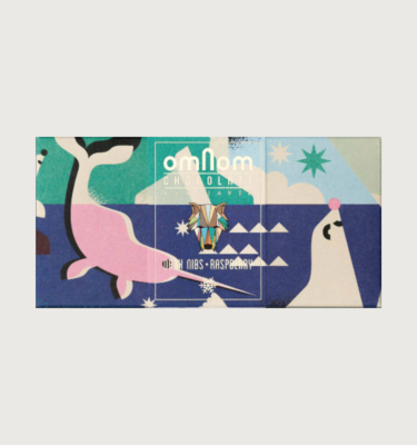 Omnom's dark nibs and raspberry bar on a grey background. The packaging is navy blue, with teal, light blue, and grey details. There is a light pink narwhal swimming in the navy blue section