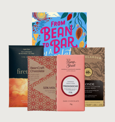 Picture of andrew baker's bean to bar gift box with chocolate bars. The book is blue, with pink and white writing, and illustrated cocoa pods on the side. There are four chocolate bars arranged in front of andrew's book.