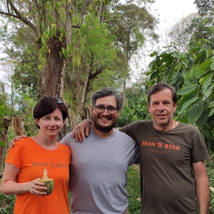 A white woman, a white man, and a mexican man smiling at the camera with trees in the background. The women has brown hair, is holding a half opened cocoa pod, and is wearing a bright orange tshirt. The white man is wearing a grey Chococard t-shirt, and the mexican man is wearing a light grey plain tshirt. 
