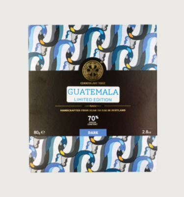 Picture of chocolate tree's dark 70% bar on a grey background. The packaging is white, light blue, dark blue, with little bits of yellow throughout. The label is a very dark brown, along the centre of the bar.