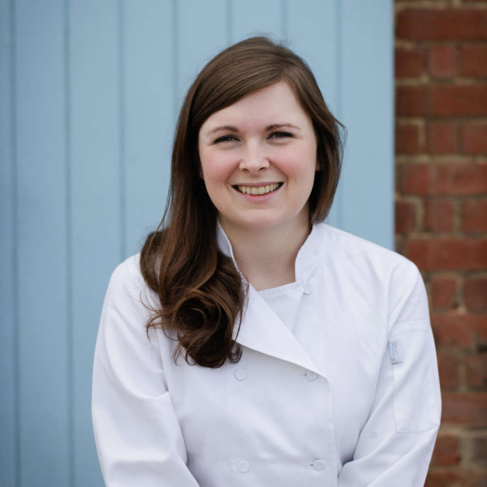 Photo of Joanne Brennan. She is wearing a chefs outfit, sitting in front of a garage door and brick wall.