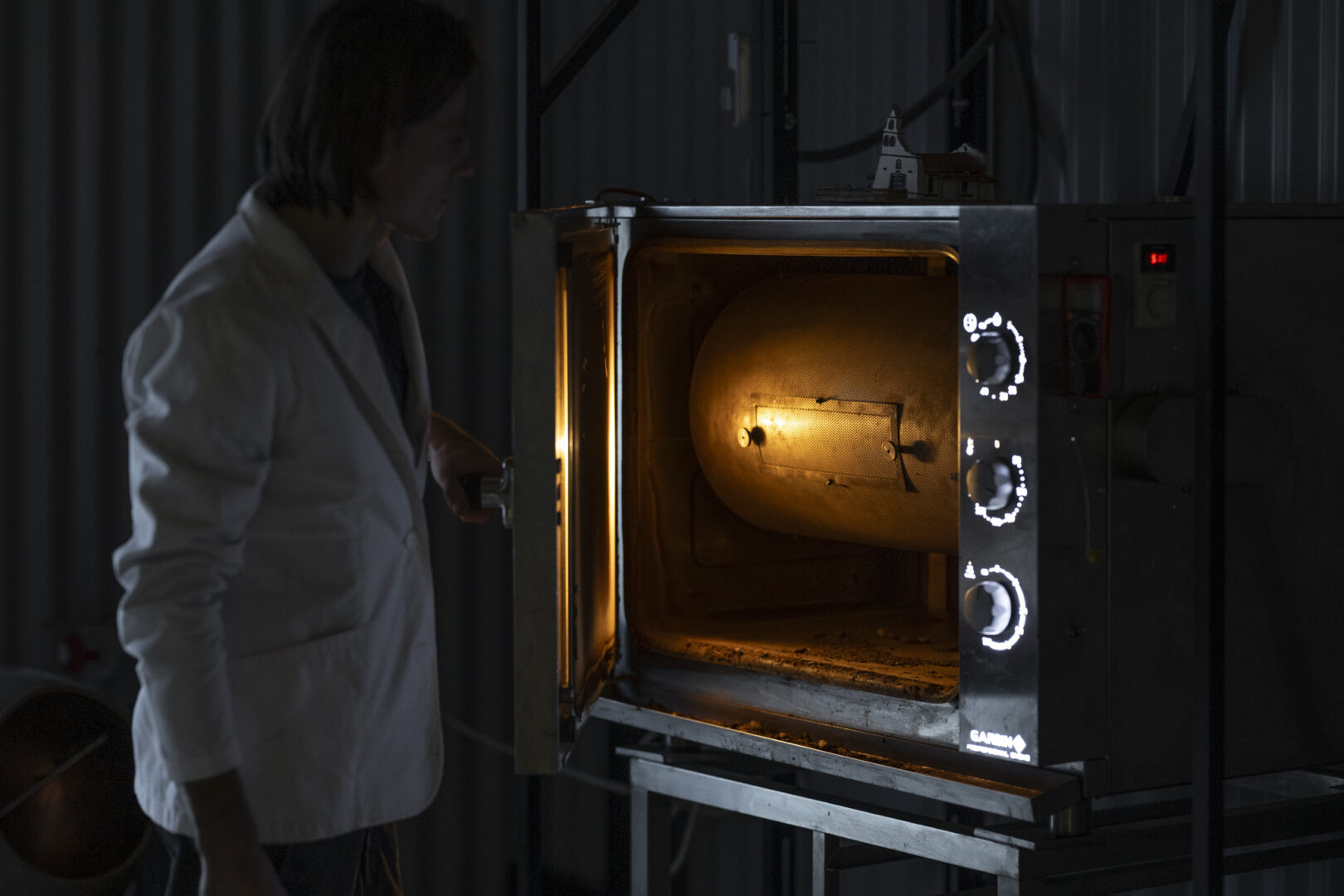 Domantas Užpalis in a lab coat stands in front of an oven. It's Dark and Moody.
