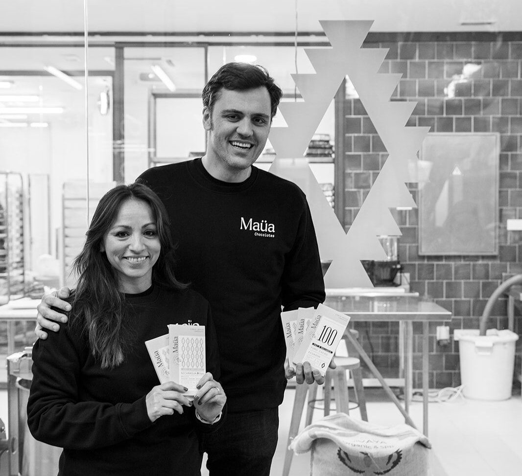 A tall man and a shorter woman grin at the camera in front of a glass shop window. The man has his hand on the woman's shoulder, and the woman is holding up two chocolate bars