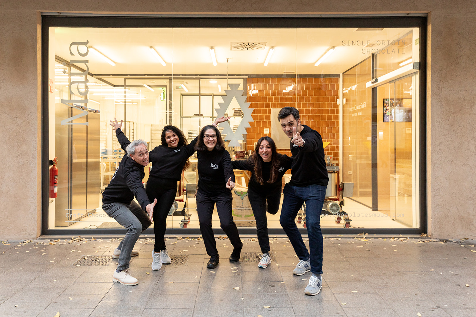 5 men and women dressed in black pose in a silly way in front of an all glass shop window, with chocolates inside