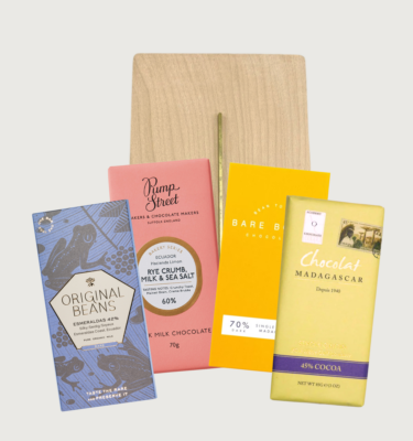 Picture of our mixed chocolate sharing board on a grey background. The sharing board is a light beige colour, and there are four bars arranged in a diamond shape in front of it.