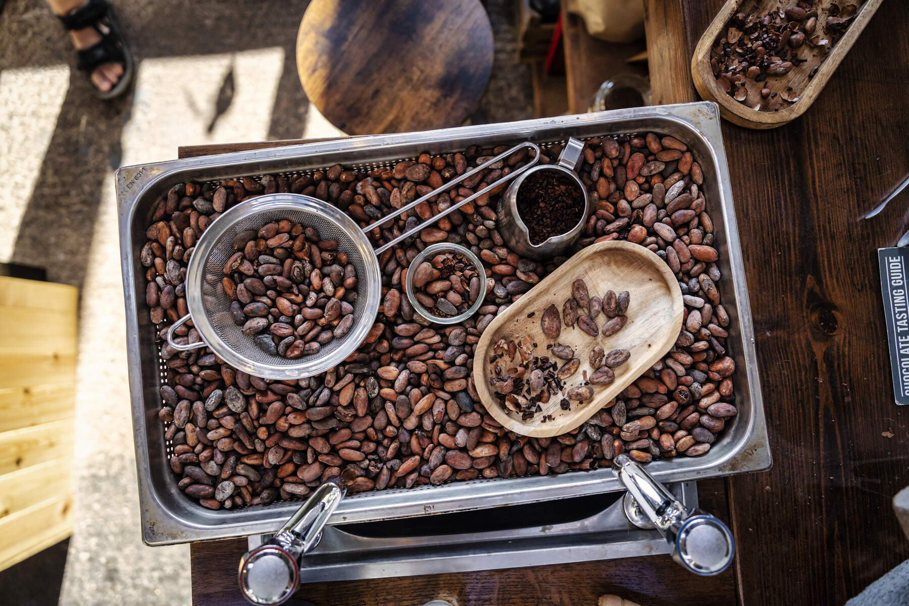 A tray of cocoa beans, with a sieve, a jug, and a plate inside.