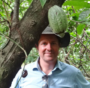 A white man in a khaki green hat and a blue button up shirt smiling behind a cocoa pod growing on a tree