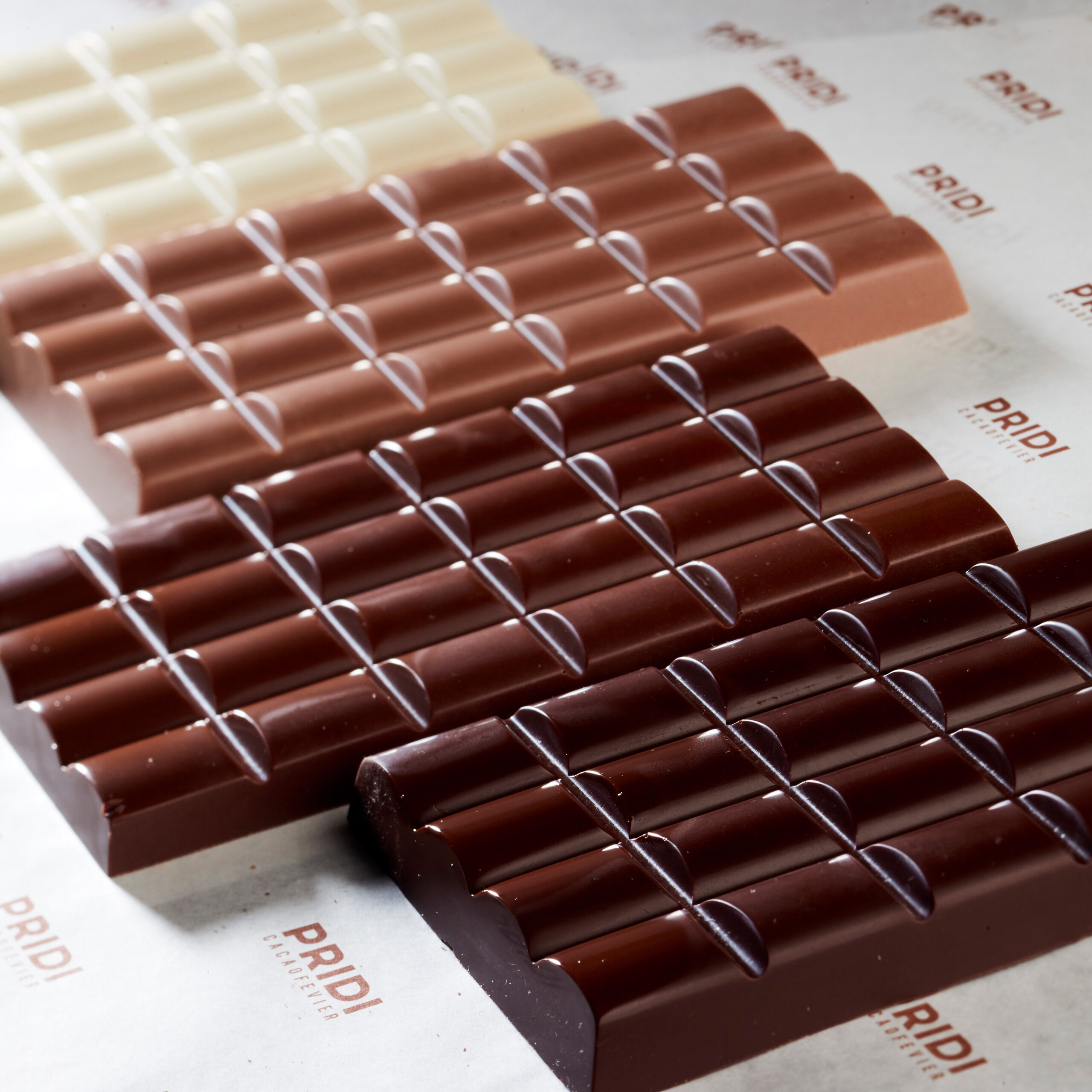 Different types of chocolate bars unwrapped and laid out on a white table with the Pridi cacaofevier logo printed all over it.