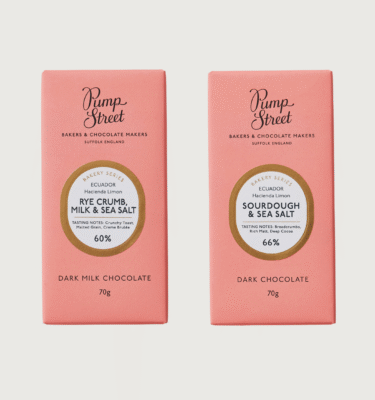 Pump street's rye and sourdough bars on a grey background. Both chocolate bars are a baby pink colour, with an oval white circle in the centre. There is a gold trim around the circle, and black writing in the centre.