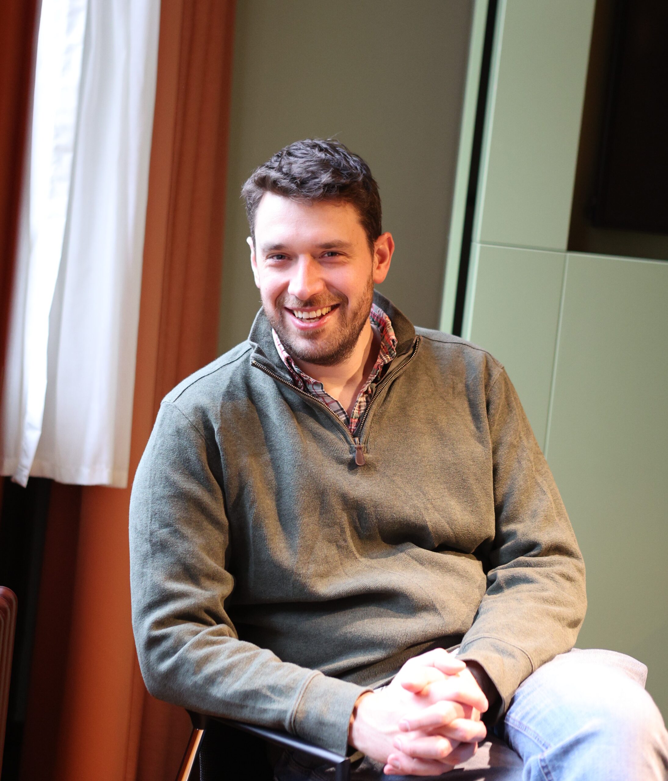 A photo of Nick Saxby, a tall white man with dark hair, sitting and smiling with his hands together.