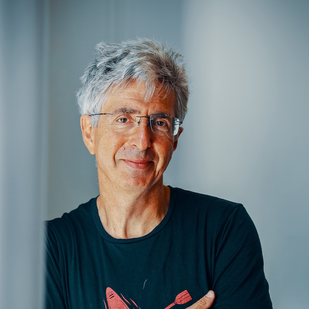 Photo of Spencer Hyman. He is wearing a t-shirt, smiling and leaning against the wall with his arms half folded.