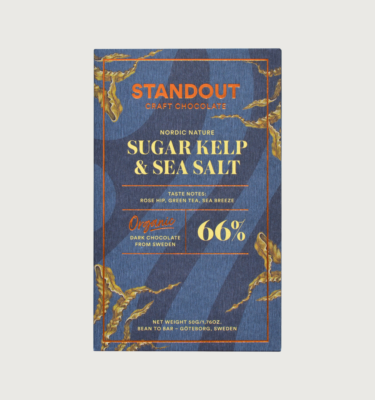 Standout's sugar kelp dark bar on a grey background. The bar is a dark greyish blue, with a darker grey swirling all over it. The writing on the bar is yellow, and the standout logo is gold and reflective. There are kelp shoots growing on the side of the bar