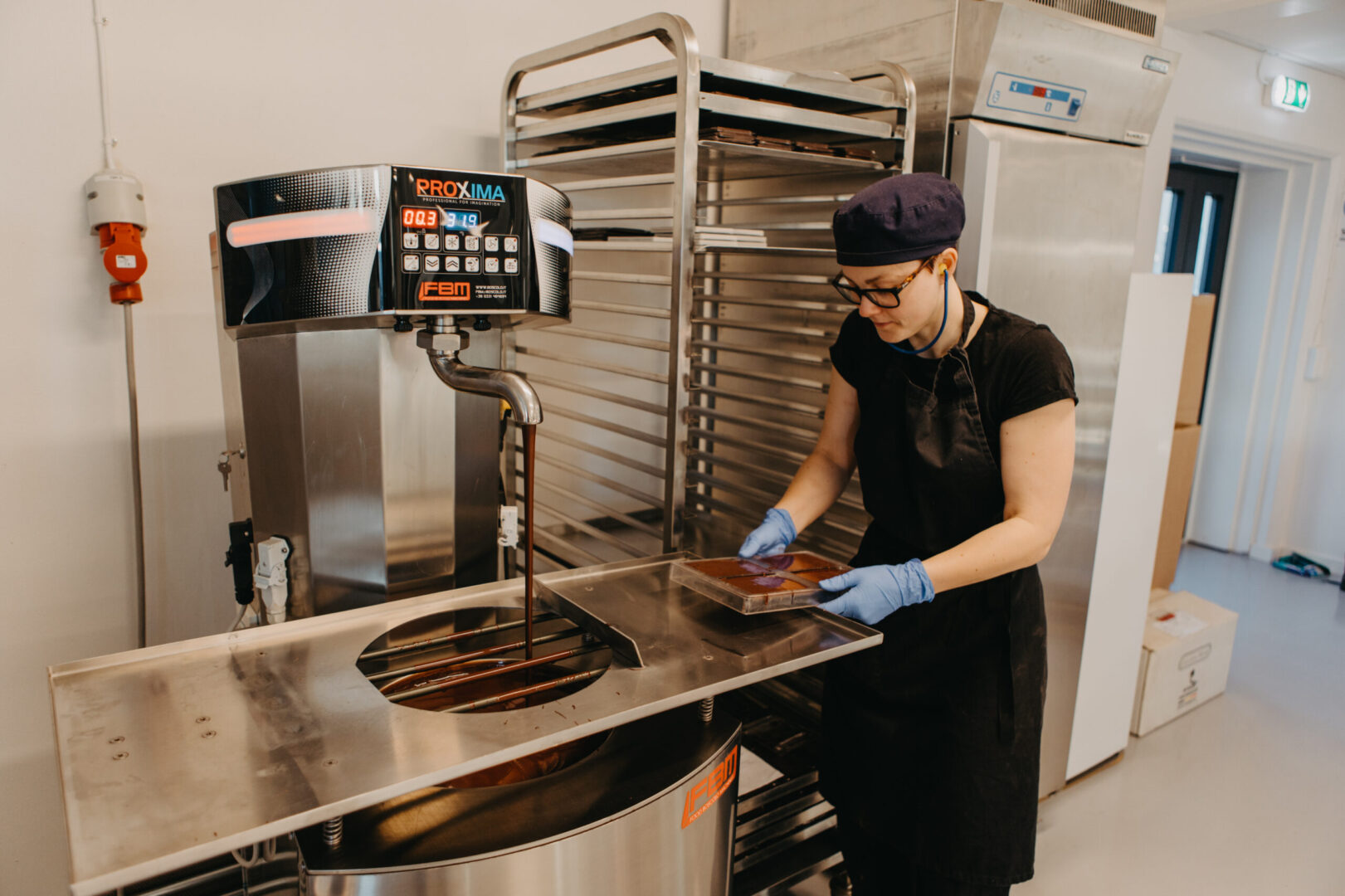 A white woman with glasses tilting a full chocolate mould on a stainless steel countertop.