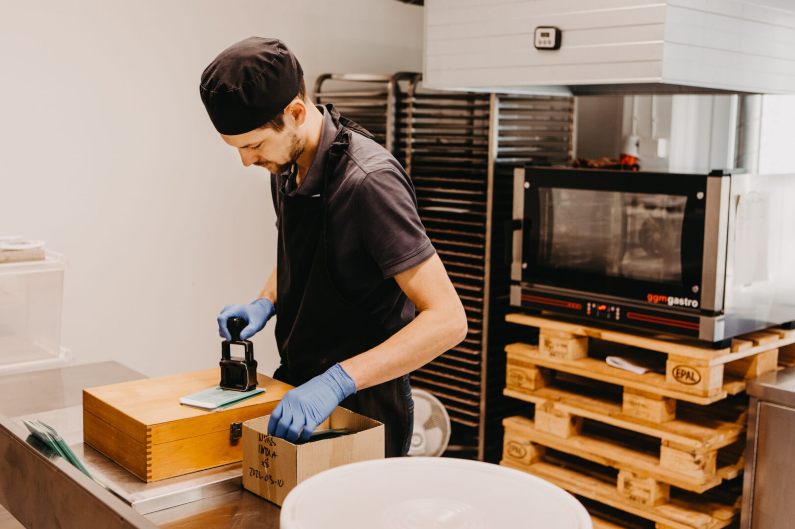 A white man stamping a chocolate bar with a date. Behind him, there is a metal tray holder, and various cabinets.