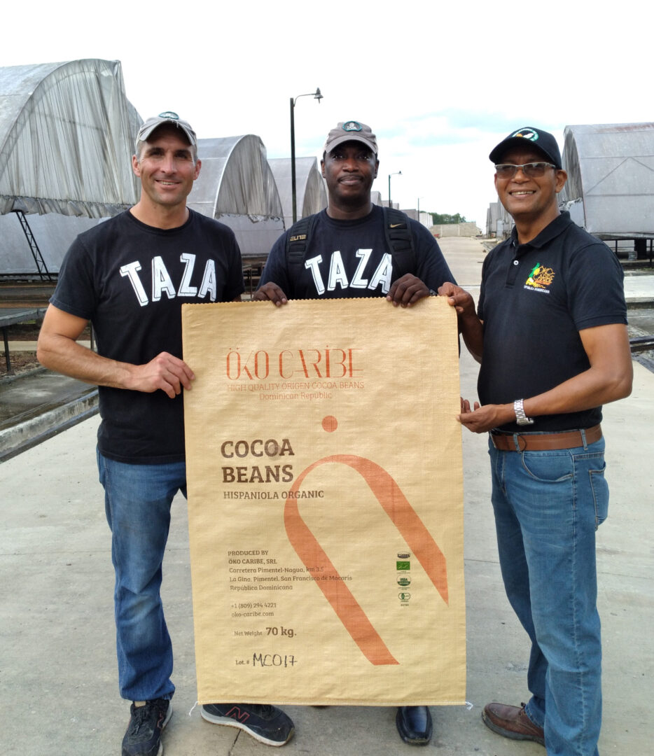 Three men standing behind a beige-coloured poster. The man in the centre and on the left are wearing black t-shirts with 'Taza' on them in white. All three men are wearing baseball caps, and smiling at the camera