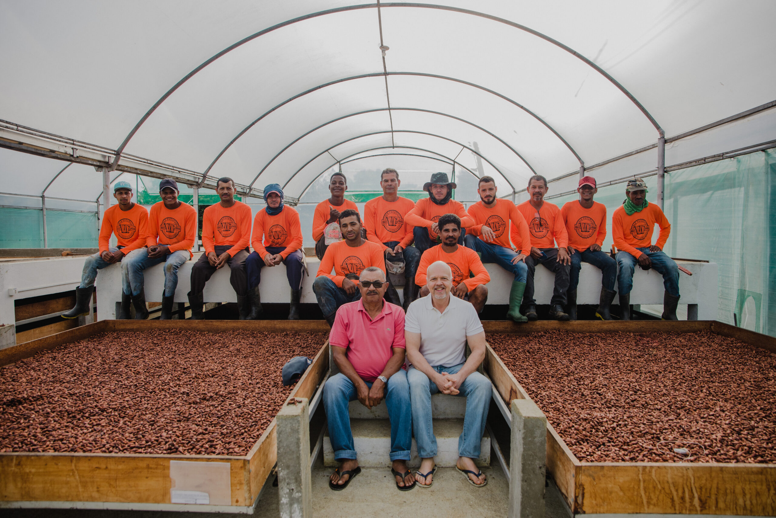 Picture of Baiani's crew in a cocoa bean processing tent. The team are wearing orange tshirts and standing up, and there are two people sitting on steps in front of them.