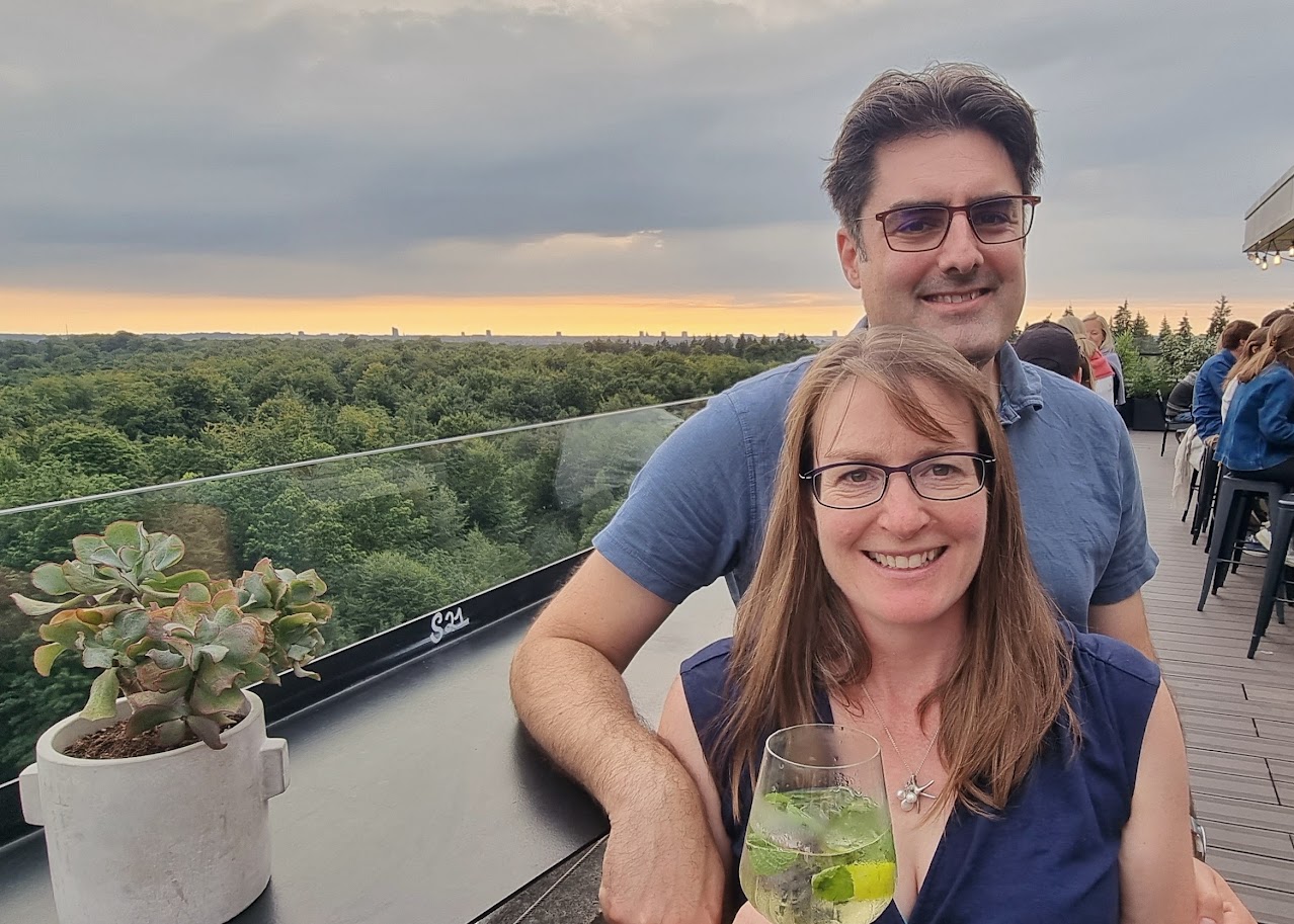 Nicky and Tom from Beancraft smiling with a drink with a lovely green view and a sunset in the background.