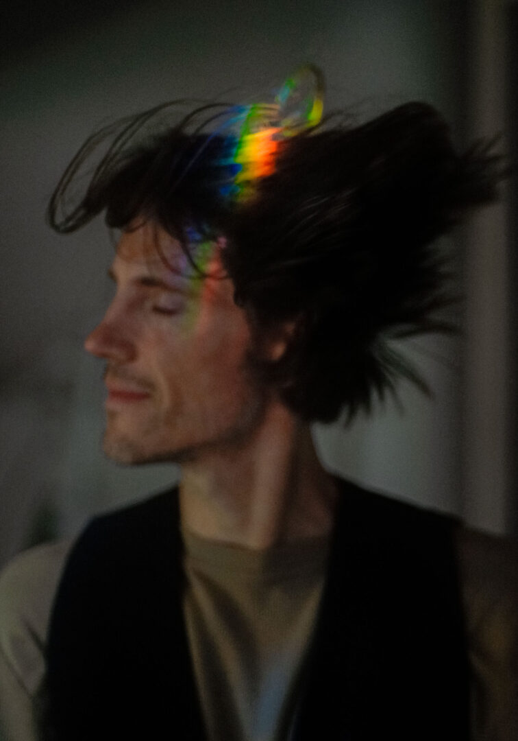 Domantas Užpalis of Naive with movement, looking over is right shoulder. There is light refracted on his head, displaying as a rainbow.
